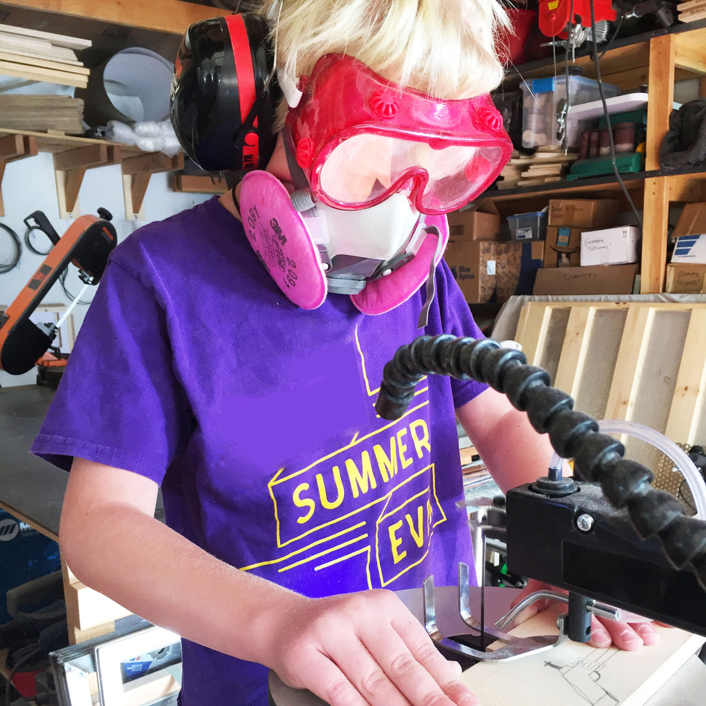 Image of a boy cutting wood on a scroll saw.  The boy is wearing red safety goggles, a respirator, and hearing protection